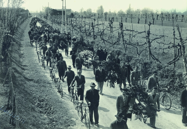 n lungo corteo funebre nelle campagne venete degli anni '50, con partecipanti a piedi e in bicicletta che portano corone di fiori.