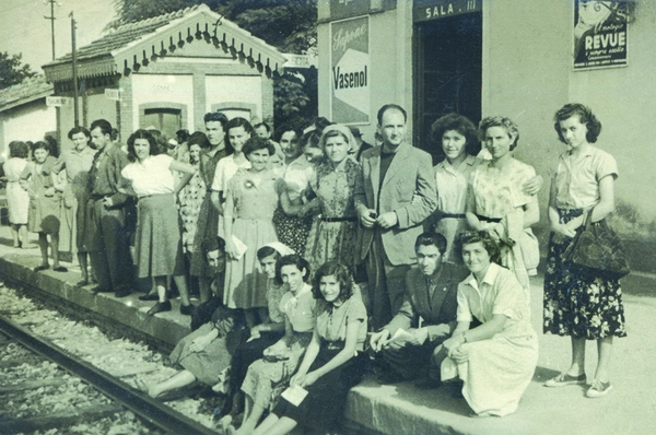Gruppo di giovani donne e ragazzi in posa alla stazione ferroviaria, probabilmente membri dell'Associazione Pionieri d'Italia negli anni '50.