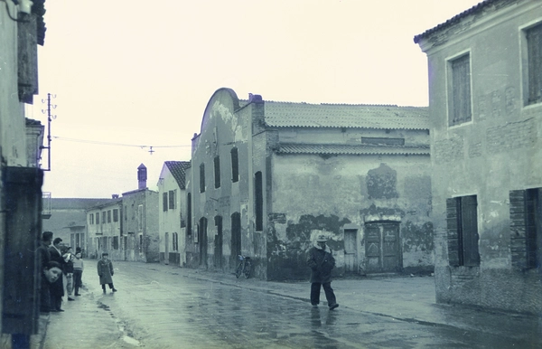 Scorcio di una strada di Pozzonovo negli anni '50 con edifici storici e poche persone in un giorno di pioggia.