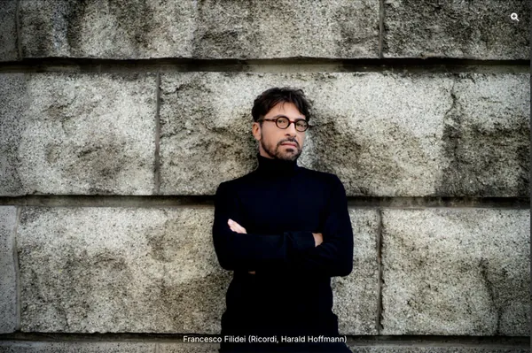 Il compositore Francesco Filidei ritratto con maglione nero a collo alto davanti a una parete di pietra